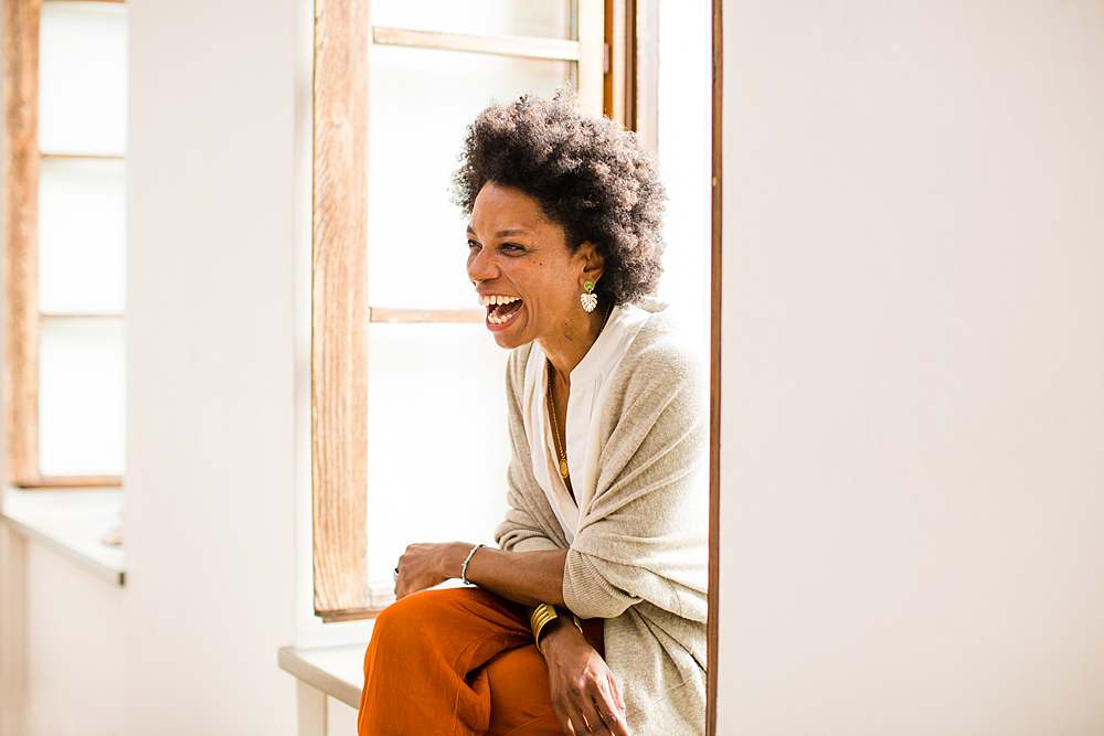 Personal-brand-photography-dark-skin-woman-laughing-sitting-by-full-length-window-in-beige-cardigan-orange-trousers