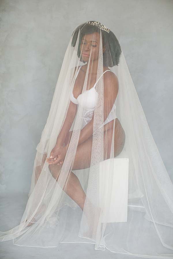 black-woman-with-white-veil-over-white-lace-corset-and-crown-sitting-down-wedding-boudoir-photo-shoot