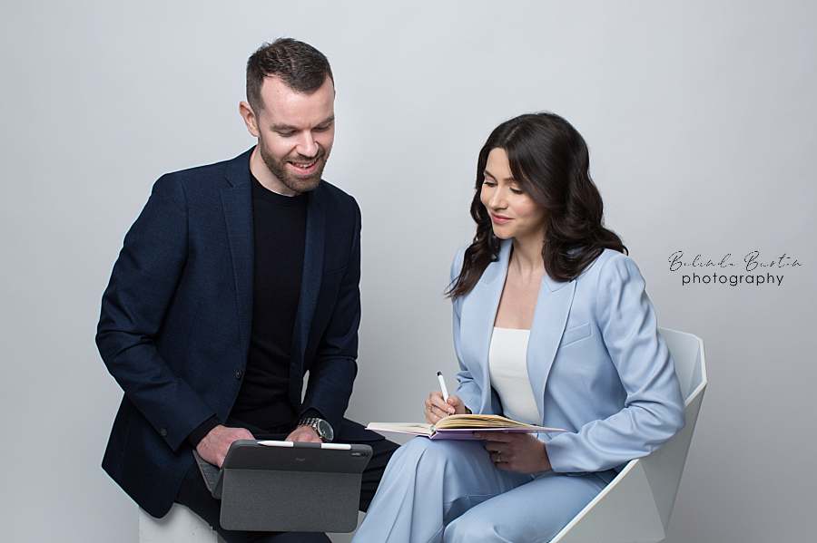 A professional team photo of a man and woman collaborating in a business setting. The man, dressed in a dark blazer and black shirt, holds a tablet, while the woman, wearing a light blue suit, takes notes in a notebook. They appear engaged and focused, conveying teamwork and productivity.