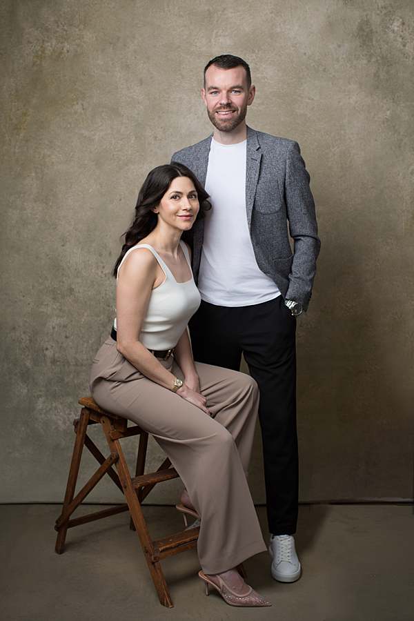 A stylish business portrait of a professional couple posing against a textured neutral backdrop. The woman is seated on a wooden stool, wearing a chic beige outfit, while the man stands beside her in a gray blazer and white t-shirt.