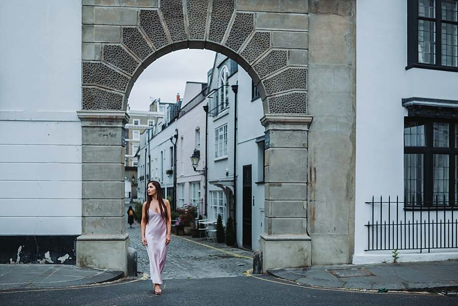 Woman in a pink dress walking under a historic archway into a cobbled mews street in London